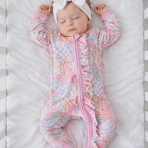 May include: A sleeping baby in a floral patterned footed onesie with a pink zip and ruffle trim. The onesie has a bow and flower design in pastel colours. The baby also wears a white bow headband.