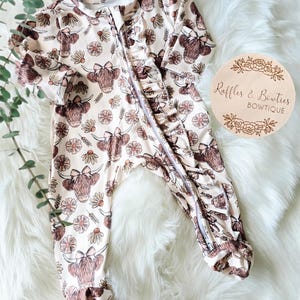 May include: A light beige baby onesie with a repeating pattern of brown Highland cow faces and daisies. The onesie has a full-length zipper, ruffled trim, and long sleeves. The brand name "Ruffles & Bowties" is on a wooden circle.