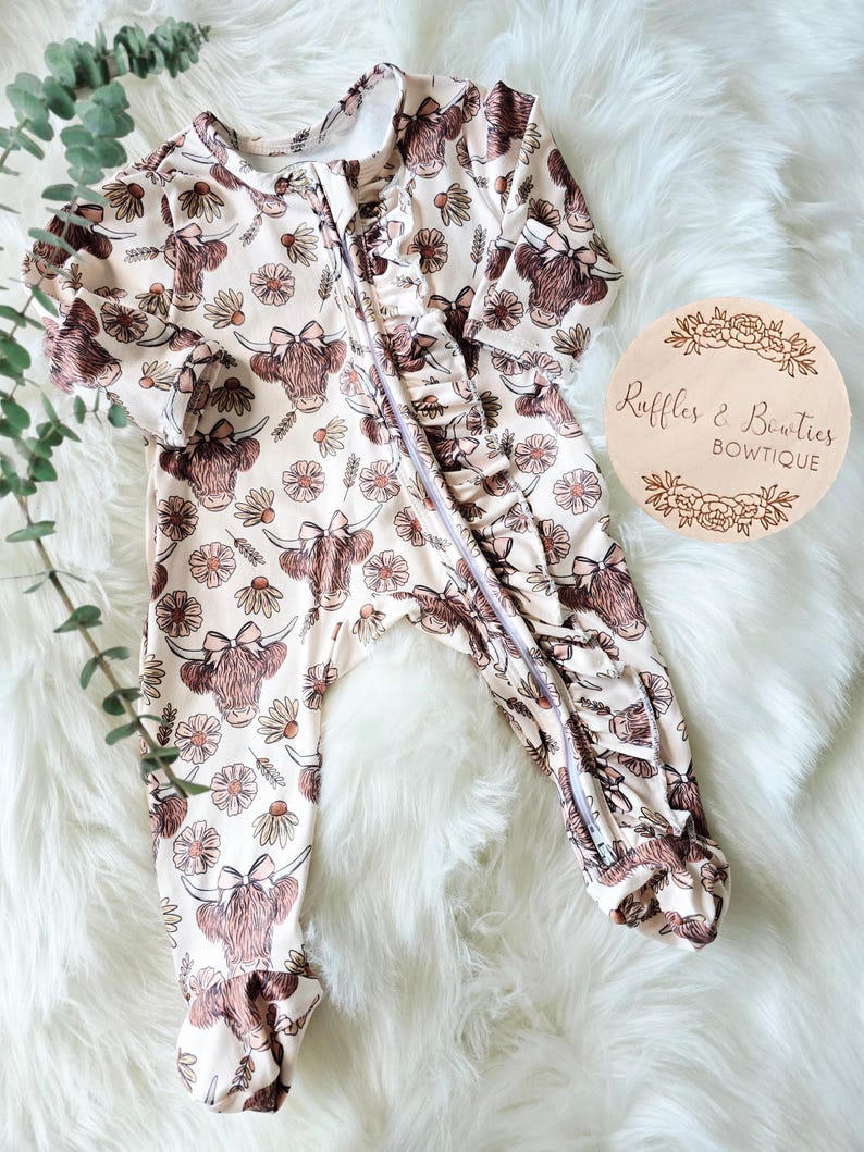May include: A light beige baby onesie with a repeating pattern of brown Highland cow faces and daisies. The onesie has a full-length zipper, ruffled trim, and long sleeves. The brand name "Ruffles & Bowties" is on a wooden circle.