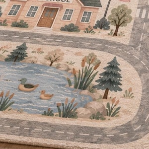May include: A rectangular area rug featuring a printed scene of a school building with the word SCHOOL above the door, an American flag, a winding gray road, and a blue pond with two ducks, surrounded by green trees and cattails on a light background.