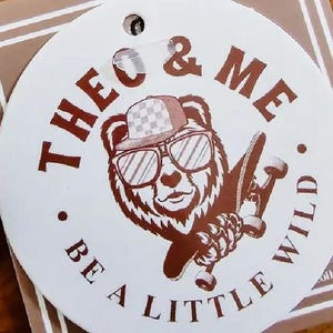 Puede incluir: Una etiqueta redonda blanca con un oso pardo con gafas de sol y un sombrero a cuadros, sosteniendo una patineta. El texto "THEO & ME" y "BE A LITTLE WILD" est&aacute; impreso en marr&oacute;n alrededor de la ilustraci&oacute;n del oso.