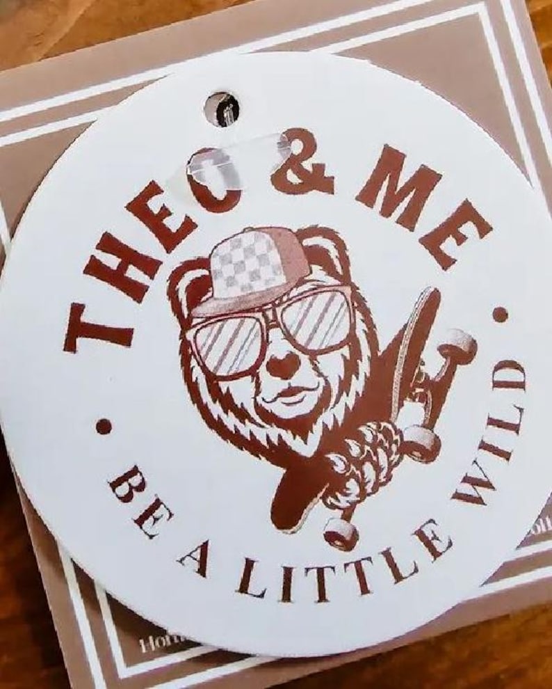 Puede incluir: Una etiqueta redonda blanca con un oso pardo con gafas de sol y un sombrero a cuadros, sosteniendo una patineta. El texto "THEO & ME" y "BE A LITTLE WILD" est&aacute; impreso en marr&oacute;n alrededor de la ilustraci&oacute;n del oso.
