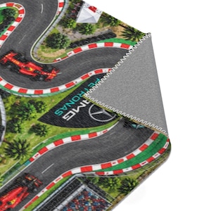 May include: A rectangular area rug featuring a printed aerial view of a Formula One racetrack with winding gray asphalt, red and white curbing, green landscaping, and red race cars. The corner is folded back to reveal a gray textured underside.