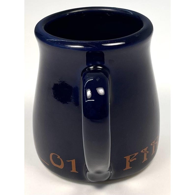 May include: A navy blue ceramic mug with a rounded body and a curved handle. The mug has a glossy finish and features the text "01 Fire" in a gold color. The mug is designed for holding hot or cold beverages.