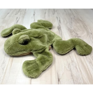 May include: A plush green frog toy with large eyes and outstretched limbs. The frog is made of soft, textured material and is lying on a light-colored wooden surface. The toy is designed to resemble a frog.
