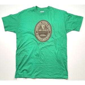 K&ouml;nnte beinhalten: Gr&uuml;nes T-Shirt mit einem braunen kreisf&ouml;rmigen Grafik des Guinness Extra Stout Logos. Das Logo zeigt den Text "Guinness Extra Stout", "St. James's Gate Dublin" und "Brewed by Guinness".