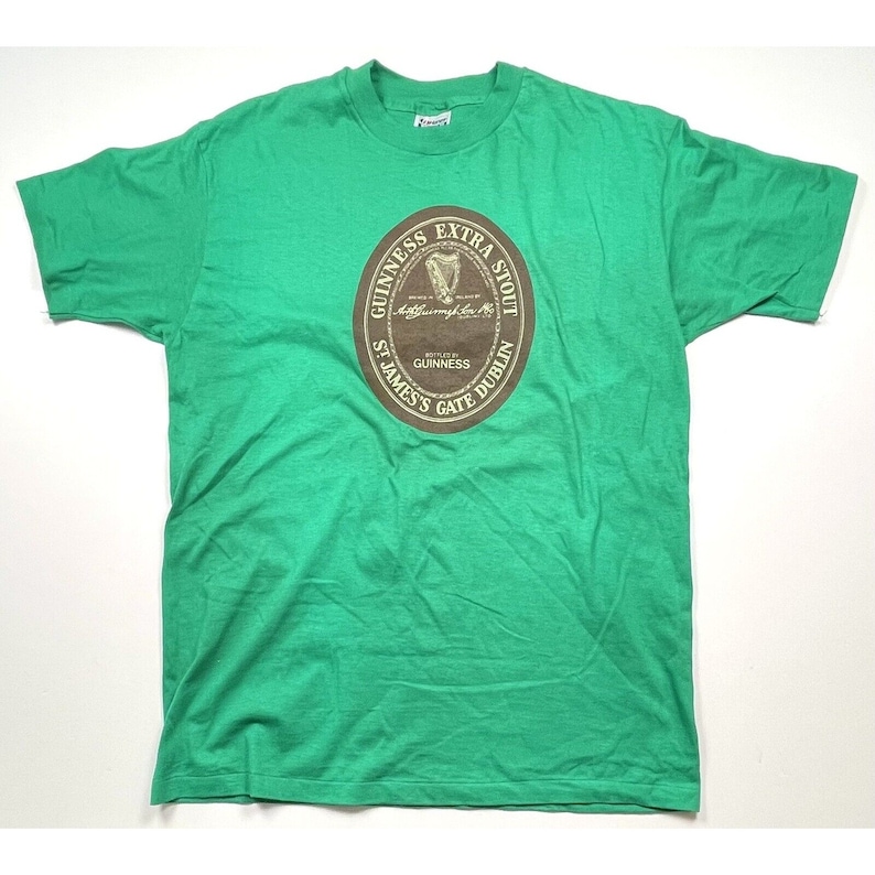 K&ouml;nnte beinhalten: Gr&uuml;nes T-Shirt mit einem braunen kreisf&ouml;rmigen Grafik des Guinness Extra Stout Logos. Das Logo zeigt den Text "Guinness Extra Stout", "St. James's Gate Dublin" und "Brewed by Guinness".