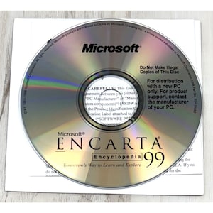 May include: A circular Microsoft Encarta 99 Encyclopedia CD-ROM with a reflective, iridescent surface. The disc features black text including the Microsoft logo, the title Encarta 99, and legal distribution warnings printed on the top half.