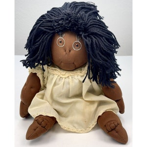 May include: A handmade rag doll with black yarn hair, brown skin, and white painted eyes. The doll is wearing a white dress with lace trim.