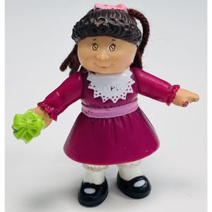 May include: A vintage Cabbage Patch Kids doll figurine. The doll has brown curly hair, a pink bow, and is wearing a magenta dress with a white collar. It holds a green bow in one hand and wears black shoes.