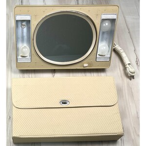 May include: A beige vanity mirror with an oval glass center, two side light panels with exposed bulbs, and starburst accents on the frame. A power cord is attached to the side. Below sits a matching beige textured carrying case with a metal clasp.