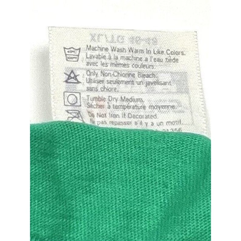 K&ouml;nnte beinhalten: Ein wei&szlig;es Pflegeetikett mit Anweisungen zum Waschen und Trocknen eines Kleidungsst&uuml;cks. Das Etikett lautet "Machine Wash Warm in Like Colors", "Lavable a la machine a l'eau ti&egrave;de avec les m&ecirc;mes couleurs", "Only Non-Chlorine Bleach", "Utiliser seulement un javelisant sans chlore", "Tumble Dry Medium", "S&eacute;cher &agrave; temp&eacute;rature moyenne", "Do Not Iron If Decorated", "Ne pas repasser s'il y a un motif".