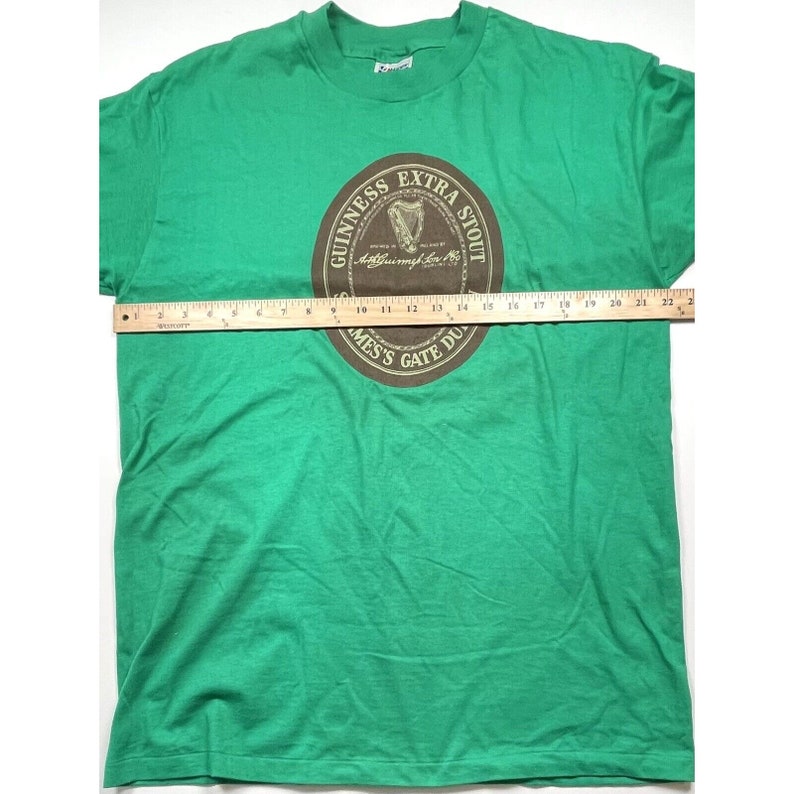 K&ouml;nnte beinhalten: Gr&uuml;nes T-Shirt mit einem braunen Kreis-Motiv, auf dem "Guinness Extra Stout" mit einer Harfe und dem Text "Arthur Guinness Son & Co. Ltd. Dublin" im Kreis steht.