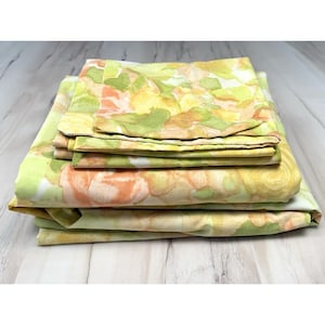 May include: A stack of folded bed sheets featuring a watercolor-style floral pattern in shades of soft peach, muted yellow, and light green. The fabric displays a repeating botanical print with blended edges, creating a layered appearance.