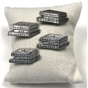 May include: Four silver-toned metal pins, each shaped like a stack of two books. The top book spine is engraved with the text 'Readers' and the bottom book spine is engraved with the text 'are Leaders'. Each pin features a small leaf icon.