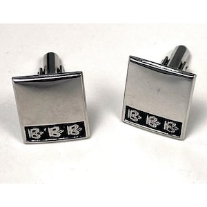 May include: A pair of silver-toned square cufflinks. Each cufflink has a black rectangular accent with the letters "B B B" in a repeating pattern. The cufflinks are on a white background.
