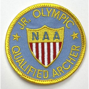 May include: A blue and yellow embroidered patch with the text "Jr. Olympic Qualified Archer" and a shield with the letters "NAA" and red and white stripes.