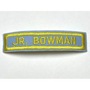 May include: A light blue fabric patch with the embroidered text "JR. BOWMAN" in gold. The text is centered within a gold border. The patch has a slightly curved shape.