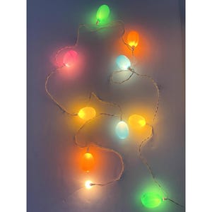 May include: A string of decorative lights featuring multiple egg-shaped bulbs in vibrant colors including green, orange, pink, blue, and yellow. The bulbs are connected by a thin white wire, and each individual bulb emits a soft, glowing light.