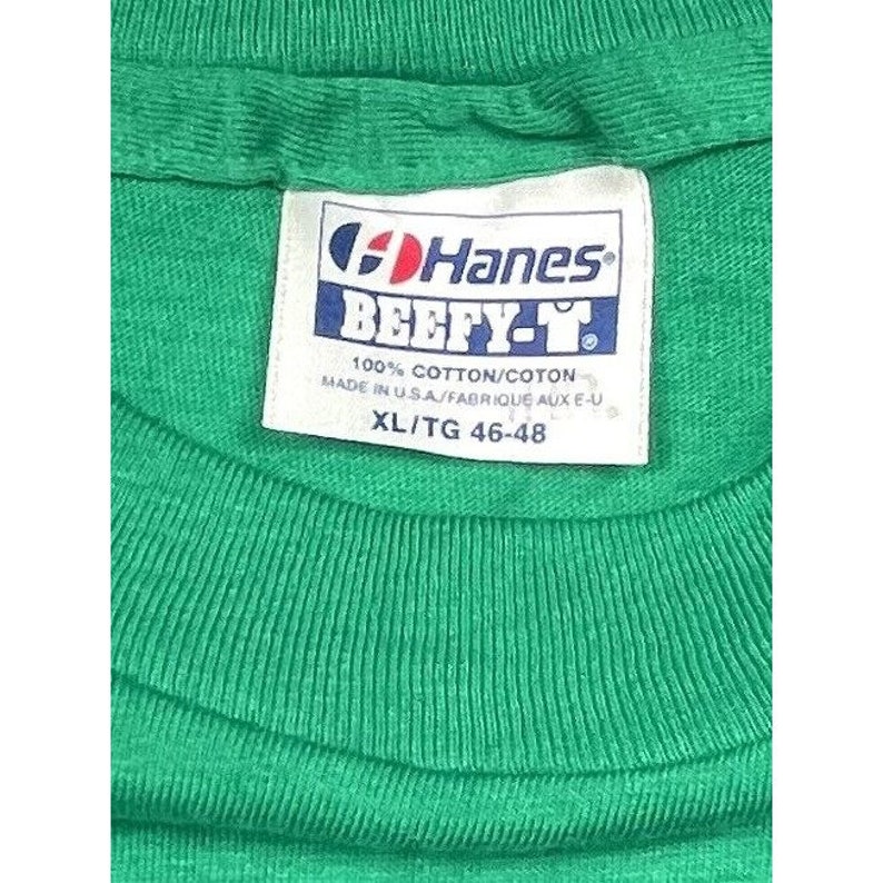 K&ouml;nnte beinhalten: Ein gr&uuml;nes Hanes Beefy-T T-Shirt mit einem wei&szlig;en Etikett, auf dem steht "Hanes Beefy-T 100% Baumwolle/Coton Hergestellt in den USA/Fabriqu&eacute; aux E-U XL/TG 46-48".