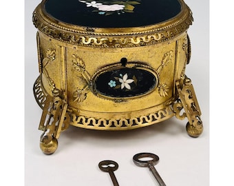 May include: A gold-toned, antique jewelry box with a black top and floral inlay. The box has ornate detailing, decorative legs, and comes with two keys. The top and front feature floral designs. The box is approximately 5 inches in diameter.