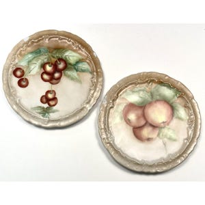 May include: Two decorative plates with hand-painted fruit designs. One plate features cherries, the other apples, both with green leaves. The plates have a gold-colored, ornate rim and a cream-colored center.