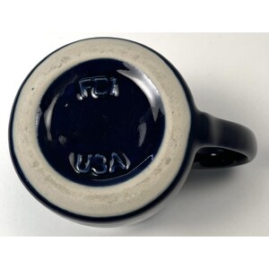 May include: A navy blue ceramic mug with a white rim and handle. The bottom of the mug features the embossed text "USA" and "FCI". The mug is viewed from the bottom.