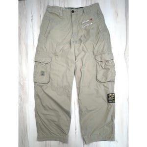 May include: Beige cargo pants featuring a button closure, two side pockets, and two large thigh cargo pockets. The pants include embroidered patches near the waist, a small square patch on the right pocket, and a rectangular JNCO brand label on the left leg.