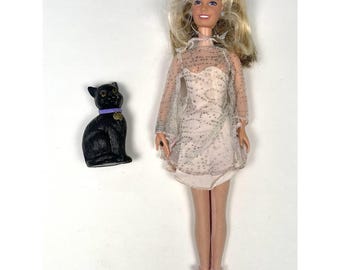 May include: A blonde doll wearing a light pink dress with a sheer overlay and pink shoes. A black cat figurine with a purple collar and gold tag is next to the doll. The doll has earrings and bracelets.