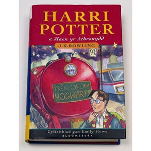 May include: A red book with the title "Harri Potter a Maen yr Athronydd" by J.K. Rowling. The cover features an illustration of a red steam train with the words "Tren Cyflym Hogwarts" on the side. The book is a Welsh translation of the first Harry Potter book.