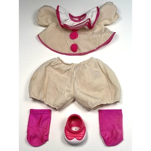 May include: A vintage doll outfit, featuring a cream-colored top with a pink-trimmed collar and two pink pom-poms. The set includes matching bloomers, pink socks, and a white and pink shoe. The outfit is designed for a doll.