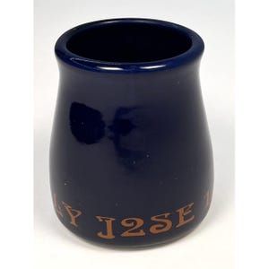 May include: A navy blue ceramic jar with a wide opening and a rounded base. The jar has a glossy finish and features the text "Y J2SE 1" in a brown, serif font around the base. The jar is likely a decorative or functional item.