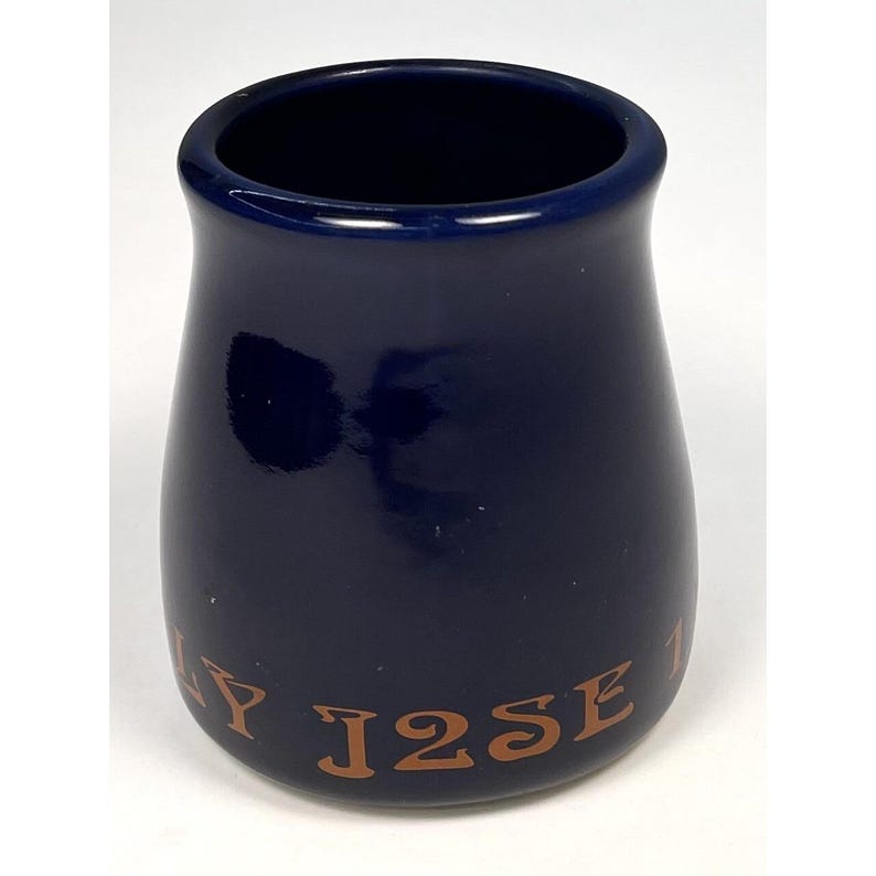 May include: A navy blue ceramic jar with a wide opening and a rounded base. The jar has a glossy finish and features the text "Y J2SE 1" in a brown, serif font around the base. The jar is likely a decorative or functional item.