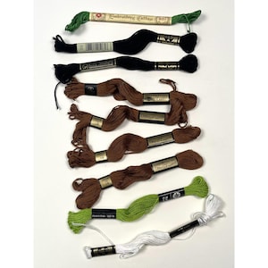 May include: A collection of embroidery floss in various colors, including green, black, brown, and white. The spools are labeled with brand names and color codes. The floss is arranged on a white surface, showcasing the different shades and textures.