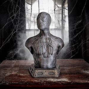 May include: A black and silver bust of Spider-Man with a web design on the chest. The bust is on a dark wooden table with a cobweb background.