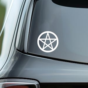 Op de afbeelding: Witte pentagramsticker, een vijfpuntige ster in een cirkel, op de achterruit van een zwarte auto. De achterlichten en het zijraam van de auto zijn ook zichtbaar. De achtergrond is wazig.