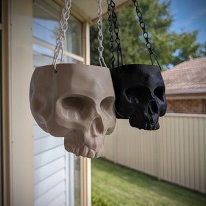 Puede incluir: Dos maceteros colgantes en forma de calavera, uno blanco y otro negro, están suspendidos de cadenas sobre un fondo borroso de una casa y un jardín.