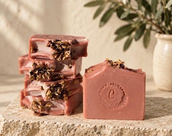 May include: A stack of four rectangular dusty rose soap bars topped with dried botanical petals, alongside a single matching bar featuring a circular embossed logo with the letter C and the text Artisan, all resting on a textured stone surface.