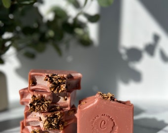 Könnte beinhalten: Ein Stapel aus vier rosafarbenen Seifenstücken mit eingebetteten getrockneten Blumen. Der oberste Seifenblock hat ein kreisförmiges Logo mit dem Buchstaben "C" und dem Wort "CHIC'S ARTISAN" darum herum.