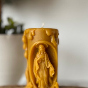 Puede incluir: Una vela de cera de abeja amarilla con una talla detallada de la Virgen María. La vela está goteada y tiene una mecha blanca.