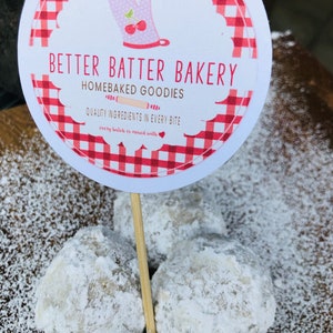 May include: A close-up of four snowballs, a type of cookie, dusted with powdered sugar. A red and white checkered sign reads "Better Batter Bakery, Homebaked Goodies, Quality Ingredients in Every Bite, Every Batch is Mixed with Love."