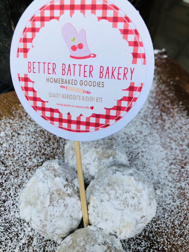May include: A close-up of four snowballs, a type of cookie, dusted with powdered sugar. A red and white checkered sign reads "Better Batter Bakery, Homebaked Goodies, Quality Ingredients in Every Bite, Every Batch is Mixed with Love."