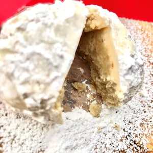 May include: A white powdered cookie with a bite taken out of it, revealing a light brown interior. The cookie is on a wooden surface covered in white powder.