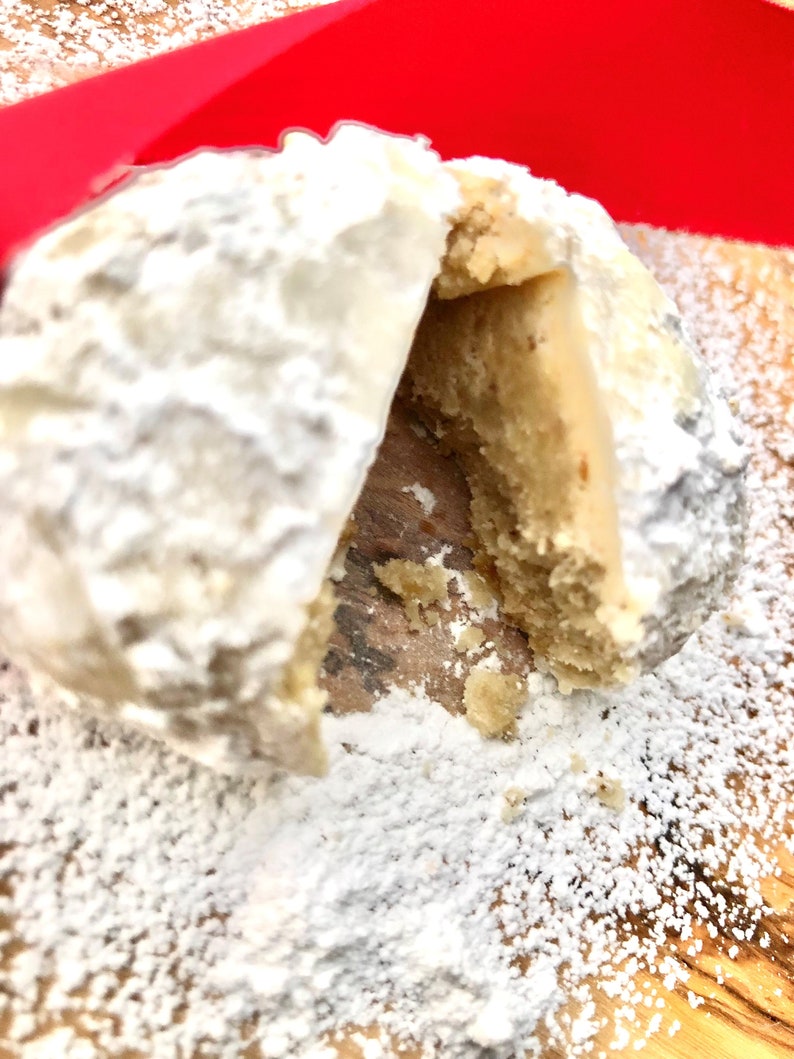 May include: A white powdered cookie with a bite taken out of it, revealing a light brown interior. The cookie is on a wooden surface covered in white powder.
