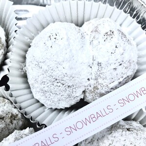 May include: A close-up of a white paper cupcake liner filled with two snowballs, a type of cookie, covered in powdered sugar. The snowballs are arranged in a heart shape. A white ribbon with pink polka dots and the text "SNOWBALLS" is visible in the foreground.