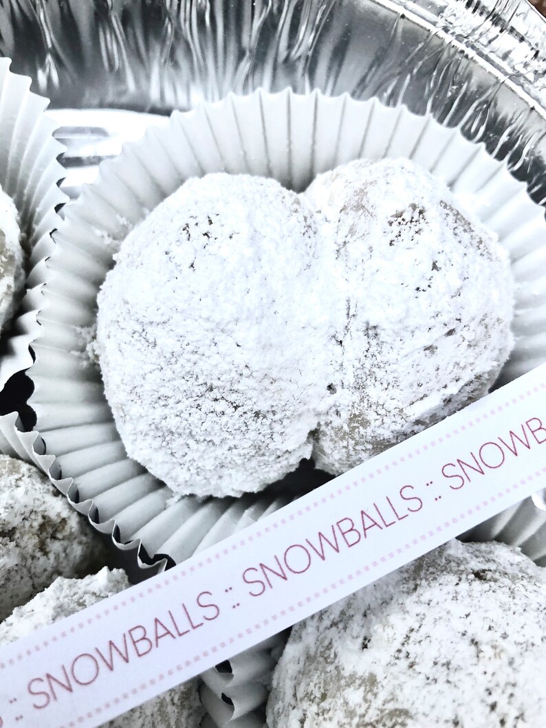 May include: A close-up of a white paper cupcake liner filled with two snowballs, a type of cookie, covered in powdered sugar. The snowballs are arranged in a heart shape. A white ribbon with pink polka dots and the text "SNOWBALLS" is visible in the foreground.