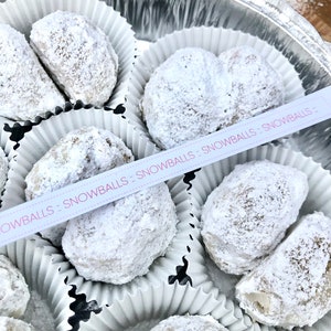 May include: A silver container filled with white paper cups containing snowballs, a type of cookie, covered in powdered sugar. The container has a pink ribbon with the text "Snowballs - Snowballs - Snowballs" on it.