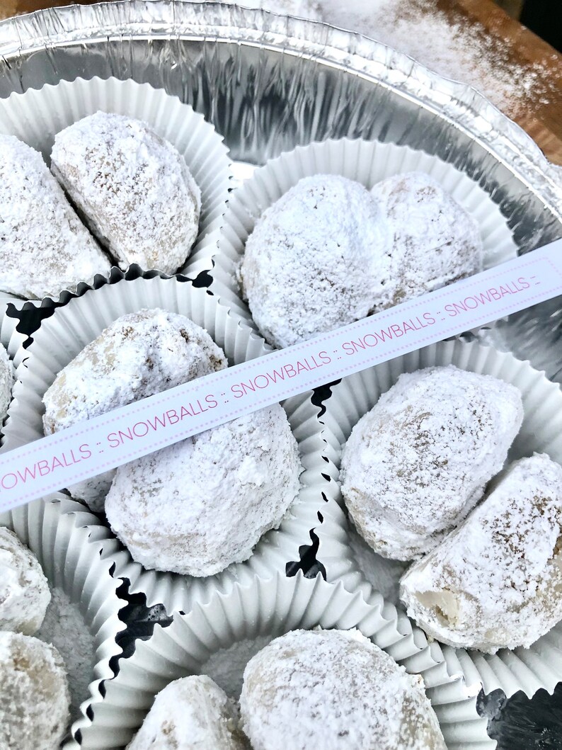 May include: A silver container filled with white paper cups containing snowballs, a type of cookie, covered in powdered sugar. The container has a pink ribbon with the text "Snowballs - Snowballs - Snowballs" on it.