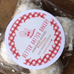 May include: A white round label with red and white checkered pattern. The label has the text "Better Batter Bakery" and "Homebaked Goodies" with the tagline "Quality Ingredients in Every Bite" and "every batch is mixed with love". The label is on a clear plastic bag with white polka dots, containing three white powdered cookies.