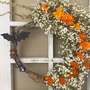 May include: A circular wreath constructed from natural twigs and wrapped with jute twine, adorned with white baby's breath, vibrant orange marigold flowers, a black bat figure, and two spiders featuring orange and purple gemstone accents.